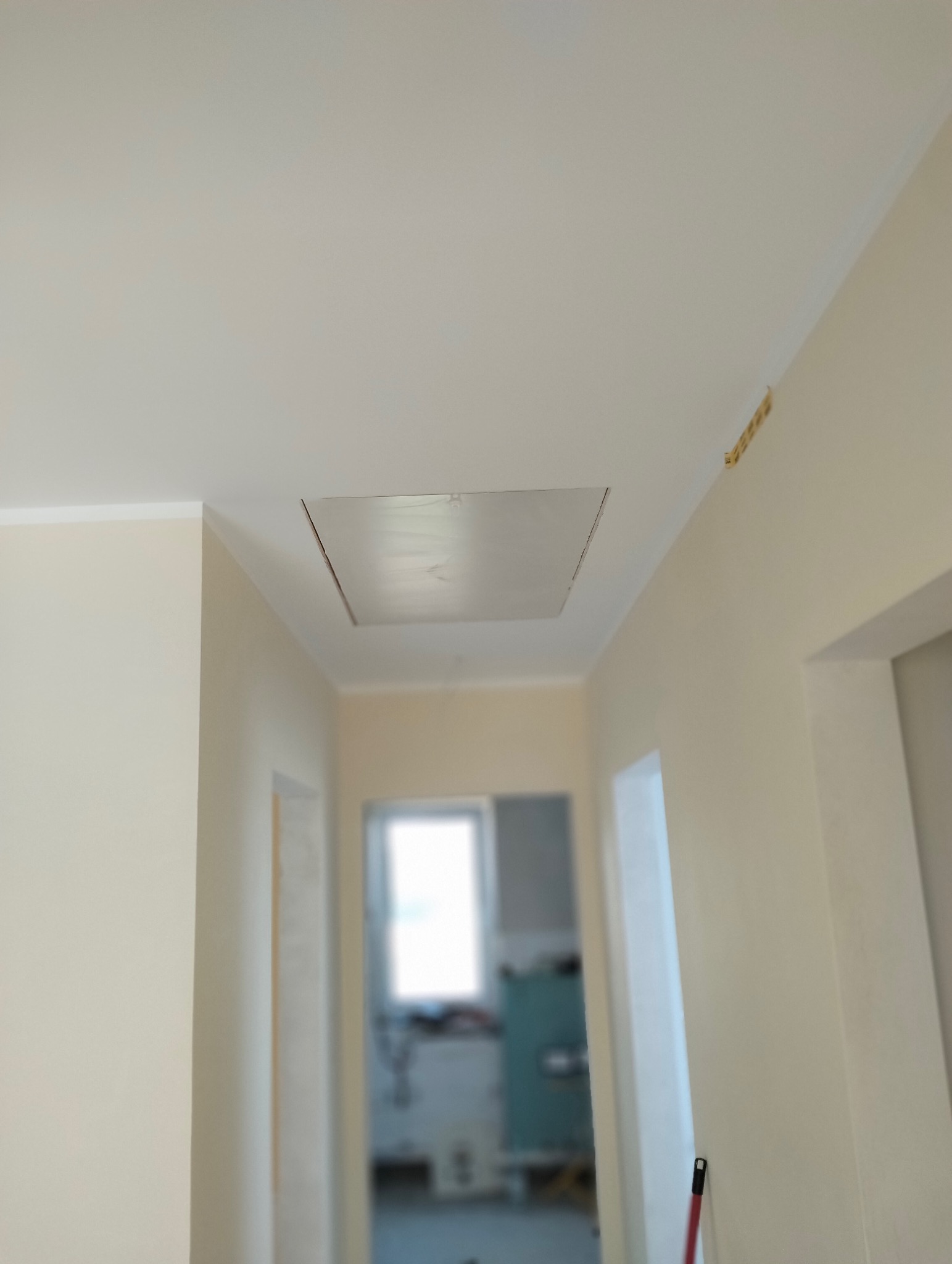 Zamknięty właz rewizyjny na suficie w korytarzu, widok z dołu. Ściany w jasnym odcieniu beżu, w tle widoczne pomieszczenie z oknem.