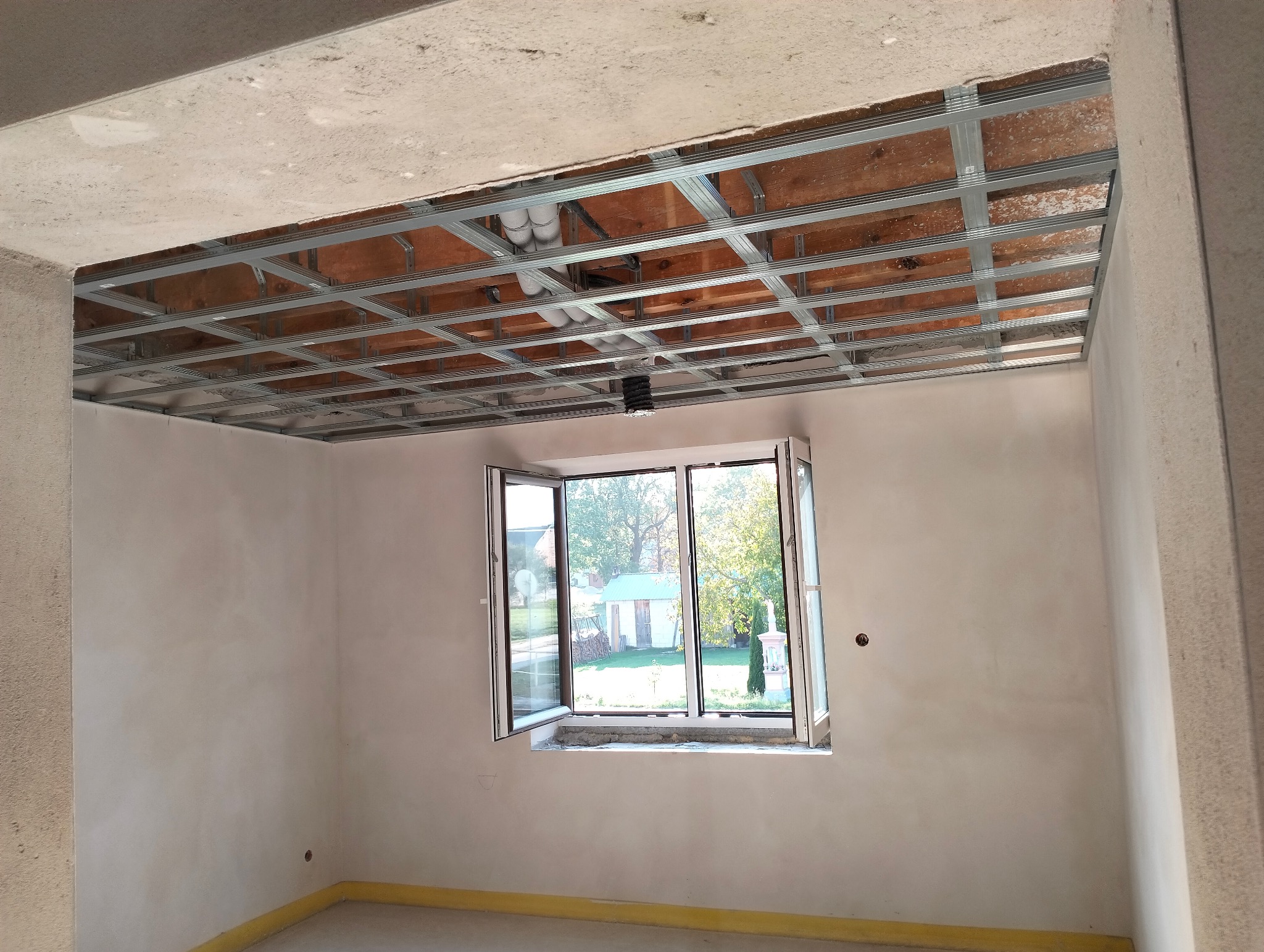 Montaż metalowej konstrukcji sufitu podwieszanego w surowym wnętrzu. Widoczne instalacje nad konstrukcją i otwarte okno z widokiem na zewnątrz.