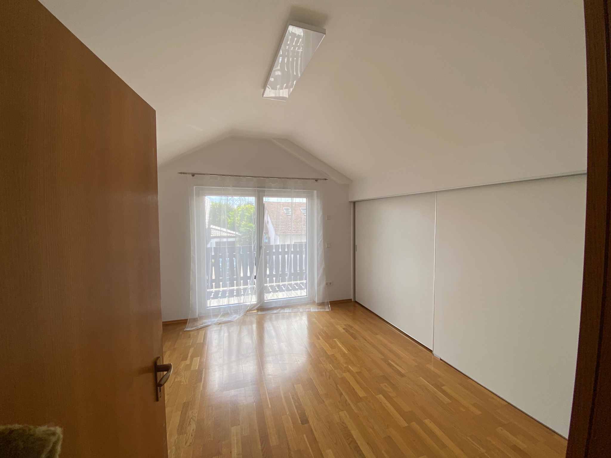 Jasne poddasze po remoncie w Gorajec-Zagroble: drewniana podłoga, białe ściany, okno balkonowe z białymi zasłonami i szafa wnękowa z przesuwnymi drzwiami.