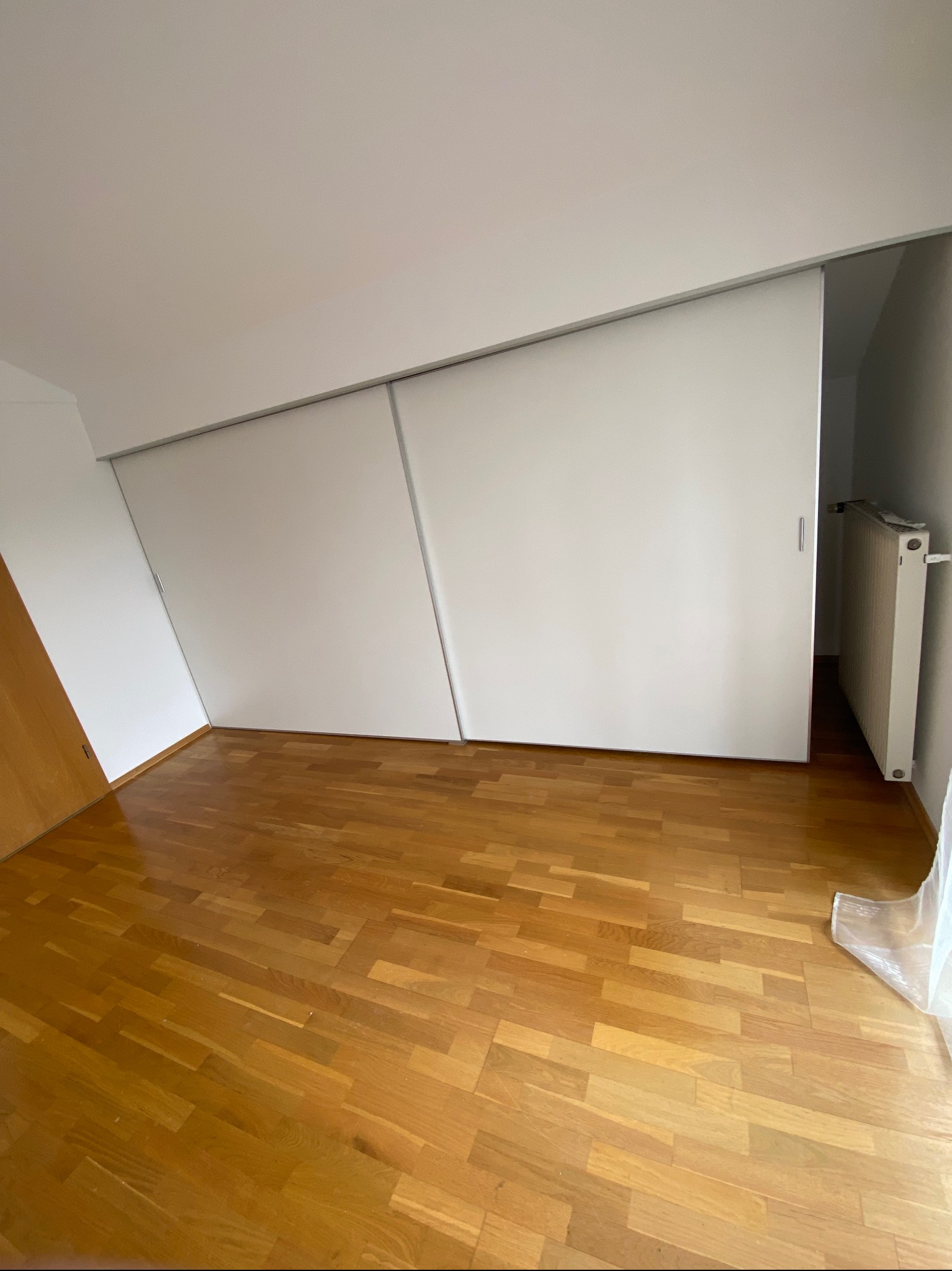 Jasne wnętrze ze skośnym sufitem i drewnianą podłogą. Widoczne białe, przesuwne drzwi szafy wnękowej z metalowymi uchwytami. Grzejnik za drzwiami.