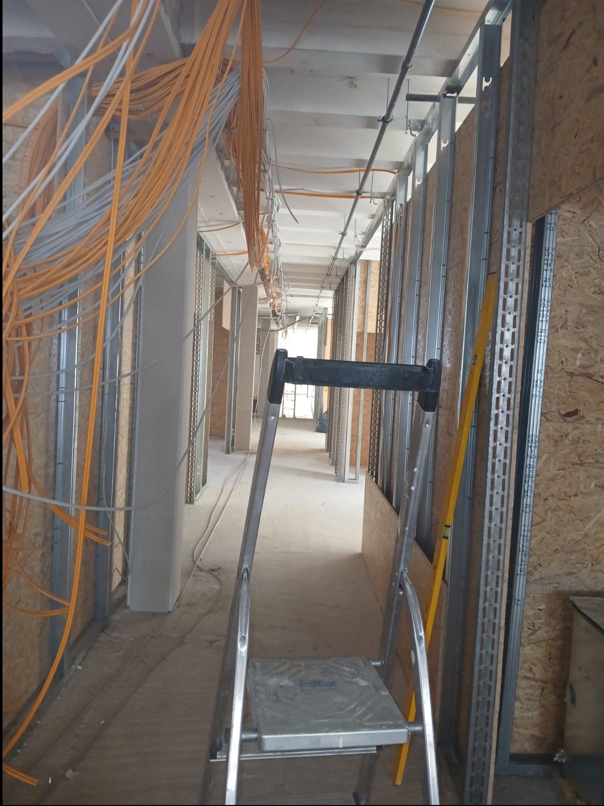 Korytarz w trakcie prac wykończeniowych: widoczne kable, metalowe profile ścian działowych, płyty OSB, drabina i poziomica. Surowy stan, przygotowanie do dalszych prac.