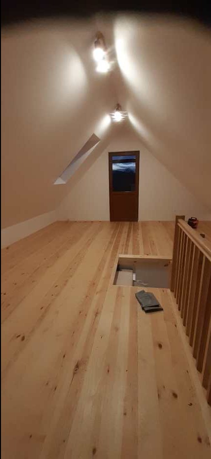 Jasne, drewniane deski podłogowe na poddaszu z oknem dachowym i drzwiami. Widoczny fragment balustrady i właz podłogowy z leżącymi obok rękawicami roboczymi.