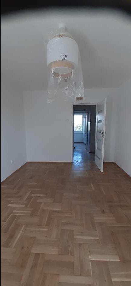 Jasne, puste wnętrze z parkietem w jodełkę. Nowa lampa w folii na suficie. Otwarte drzwi do kolejnego pomieszczenia. Świeżo wykończone mieszkanie.