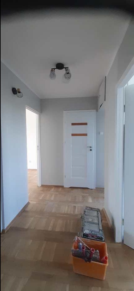 Jasny korytarz po remoncie: parkiet, białe ściany, drzwi z poziomymi paskami, nowoczesne lampy. Na podłodze narzędzia malarskie, sugerujące świeże malowanie.