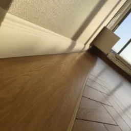 Ukończona podłoga z paneli w odcieniach brązu, z widoczną listwą przypodłogową i fragmentem okna. Ujęcie z niskiej perspektywy, podkreślające strukturę drewna.