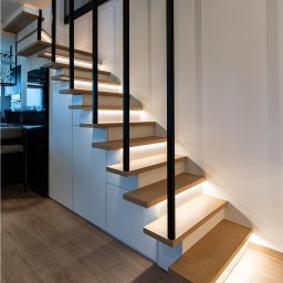 Usługi budowlane Arkadiusz komar - Nowoczesne schody drewniane z podświetleniem LED, czarne metalowe barierki, minimalistyczny design, widok z dołu, parkiet na podłodze, białe szafki pod schodami.