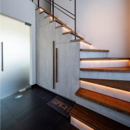 Usługi budowlane Arkadiusz komar - Nowoczesne, podświetlane schody drewniane z czarną balustradą i szarymi panelami ściennymi. Widoczny fragment czarnej podłogi i drzwi.