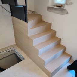 Usługi budowlane Arkadiusz komar - Montaż schodów drewnianych z metalową barierką w Daniłowie Małym. Jasne drewno, minimalistyczny design, widoczne niedokończone wykończenia ścian.