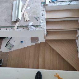 Usługi budowlane Arkadiusz komar - Montaż drewnianych schodów z jasnego drewna, widoczne narzędzia i materiały, etap wykończeniowy, ujęcie z góry, fragment buta w kadrze.
