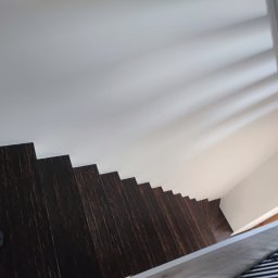 Usługi budowlane Arkadiusz komar - Ujęcie z góry na drewniane schody z ciemnego drewna, przy białej ścianie i suficie. Widoczny fragment osoby w dolnym lewym rogu.