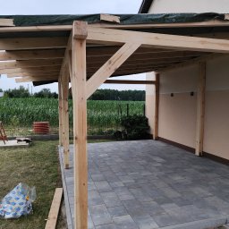 Usługi budowlane Arkadiusz komar - Drewniana pergola w trakcie budowy, z widocznymi elementami konstrukcyjnymi i częściowo wyłożoną kostką brukową. W tle pole kukurydzy i fragment budynku.