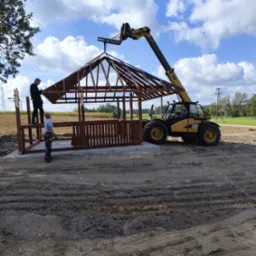 Montaż altany z drewnianą balustradą i zadaszeniem przy użyciu podnośnika CAT na tle pola i błękitnego nieba z chmurami. Dwóch monterów przy pracy.