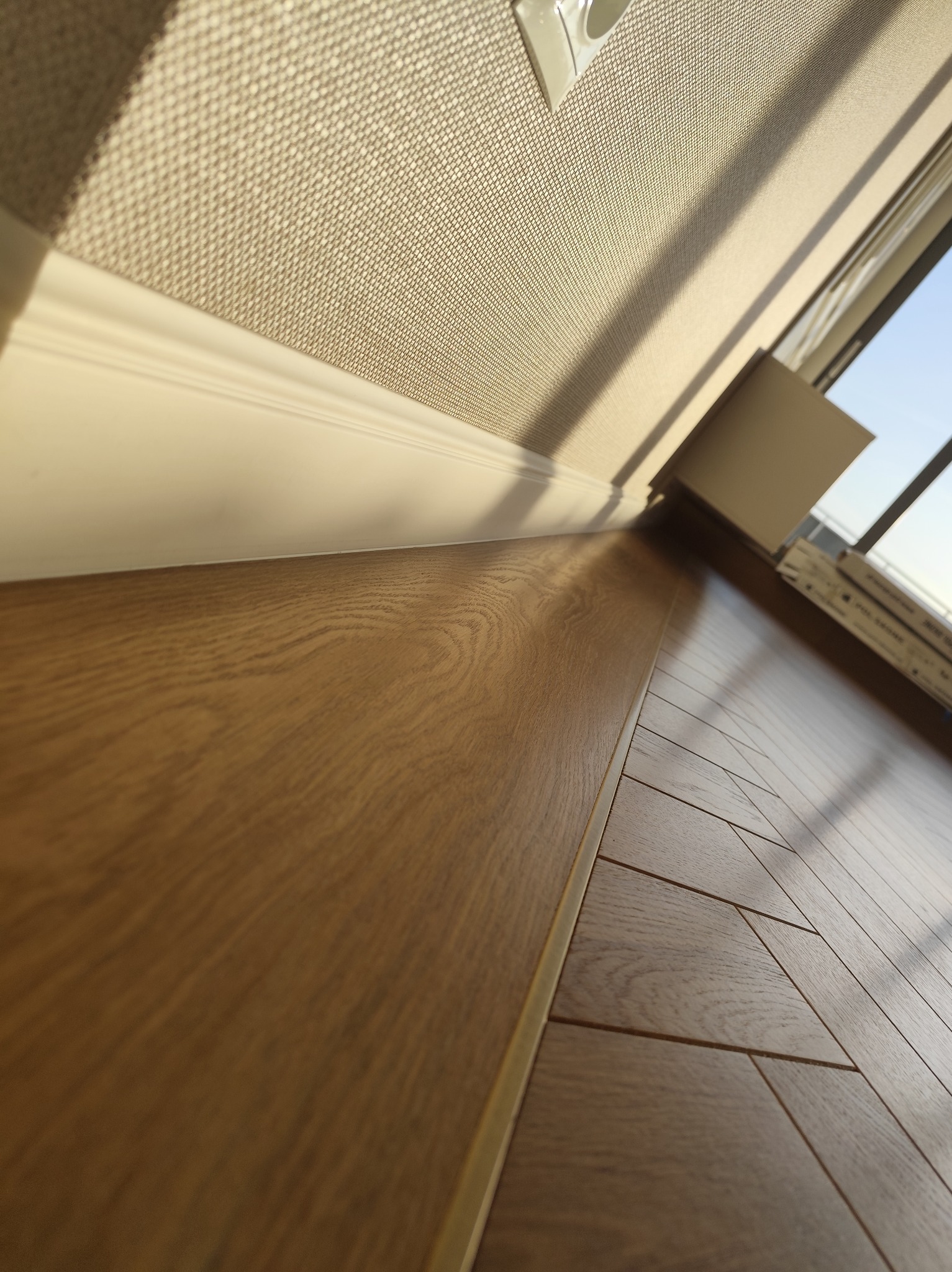 Ukończona podłoga z paneli w odcieniach brązu, z widoczną listwą przypodłogową i fragmentem okna. Ujęcie z niskiej perspektywy, podkreślające strukturę drewna.