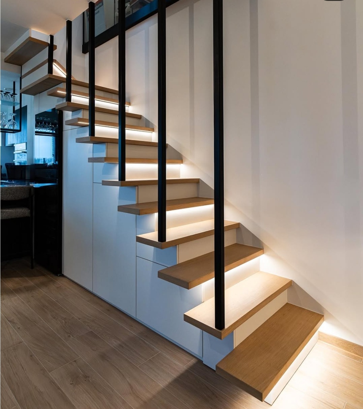 Nowoczesne schody drewniane z podświetleniem LED, czarne metalowe barierki, minimalistyczny design, widok z dołu, parkiet na podłodze, białe szafki pod schodami.