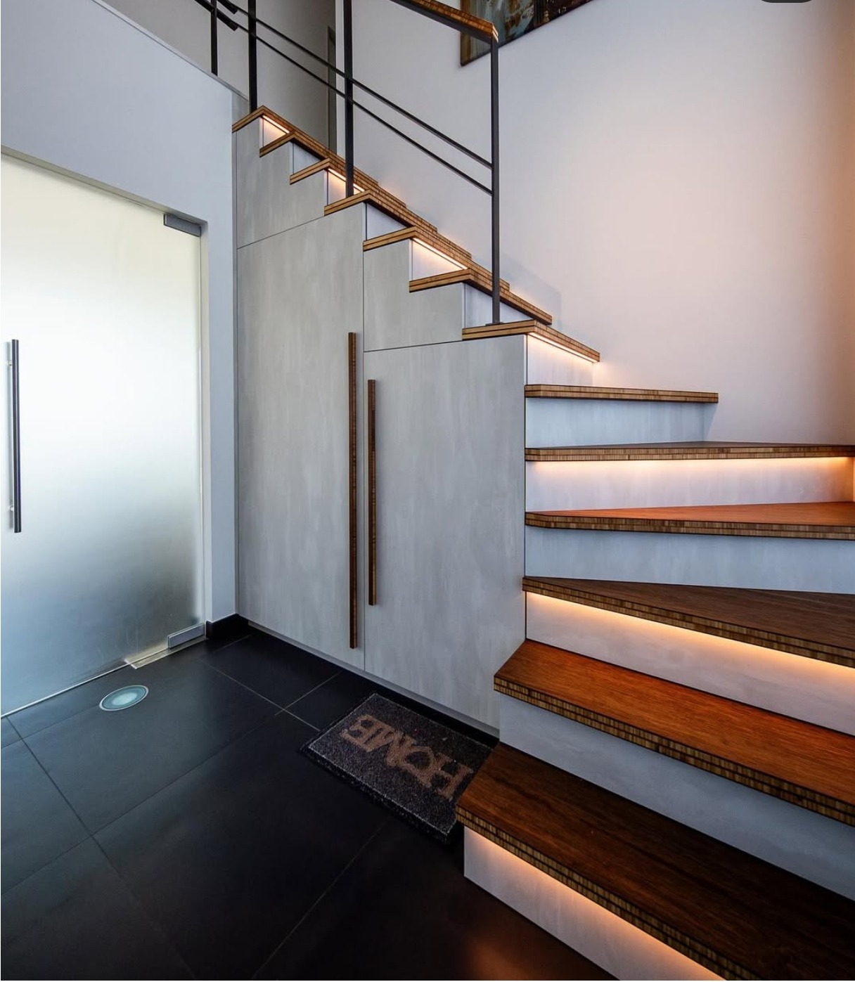 Nowoczesne, podświetlane schody drewniane z czarną balustradą i szarymi panelami ściennymi. Widoczny fragment czarnej podłogi i drzwi.
