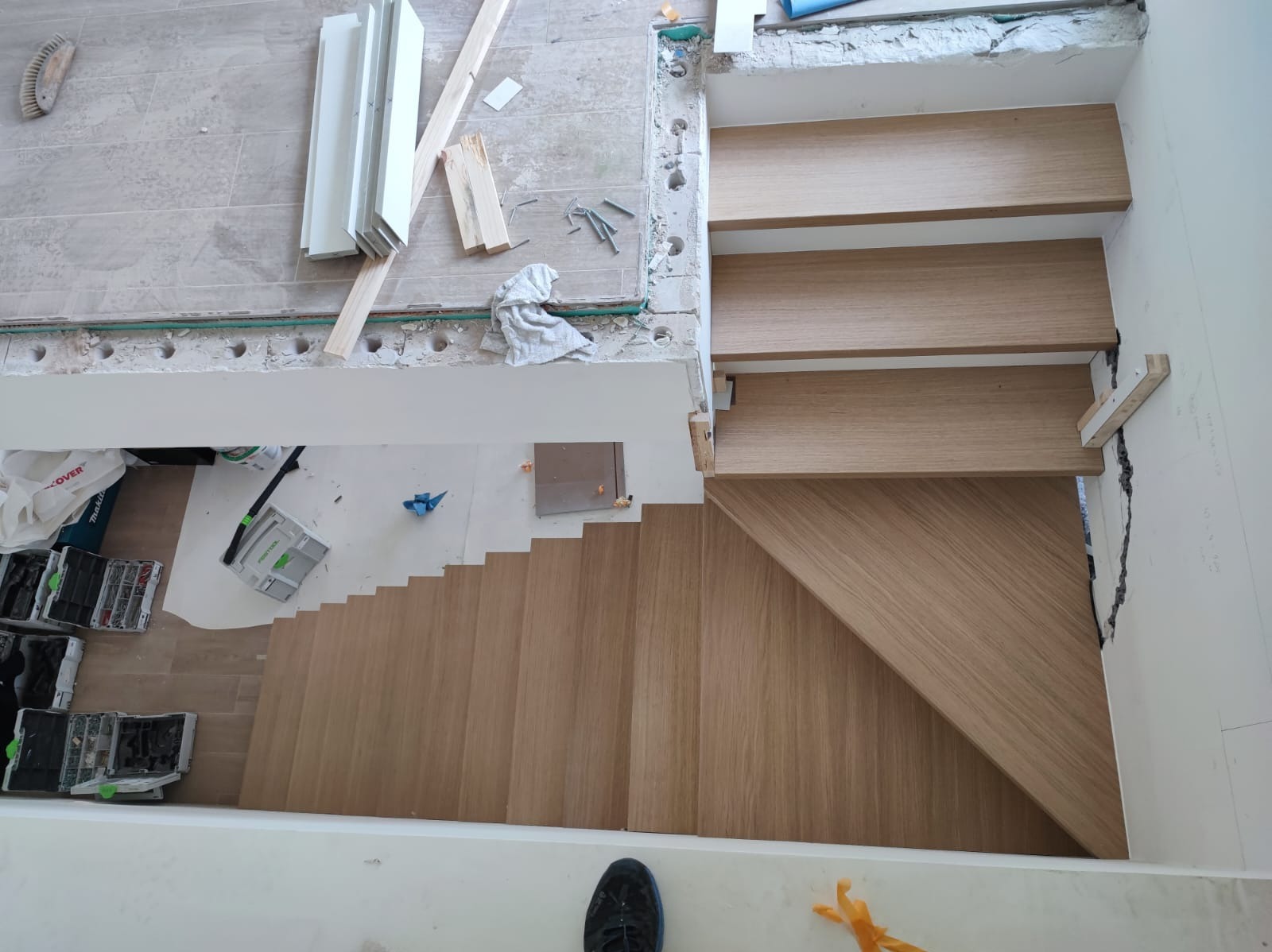 Montaż drewnianych schodów z jasnego drewna, widoczne narzędzia i materiały, etap wykończeniowy, ujęcie z góry, fragment buta w kadrze.