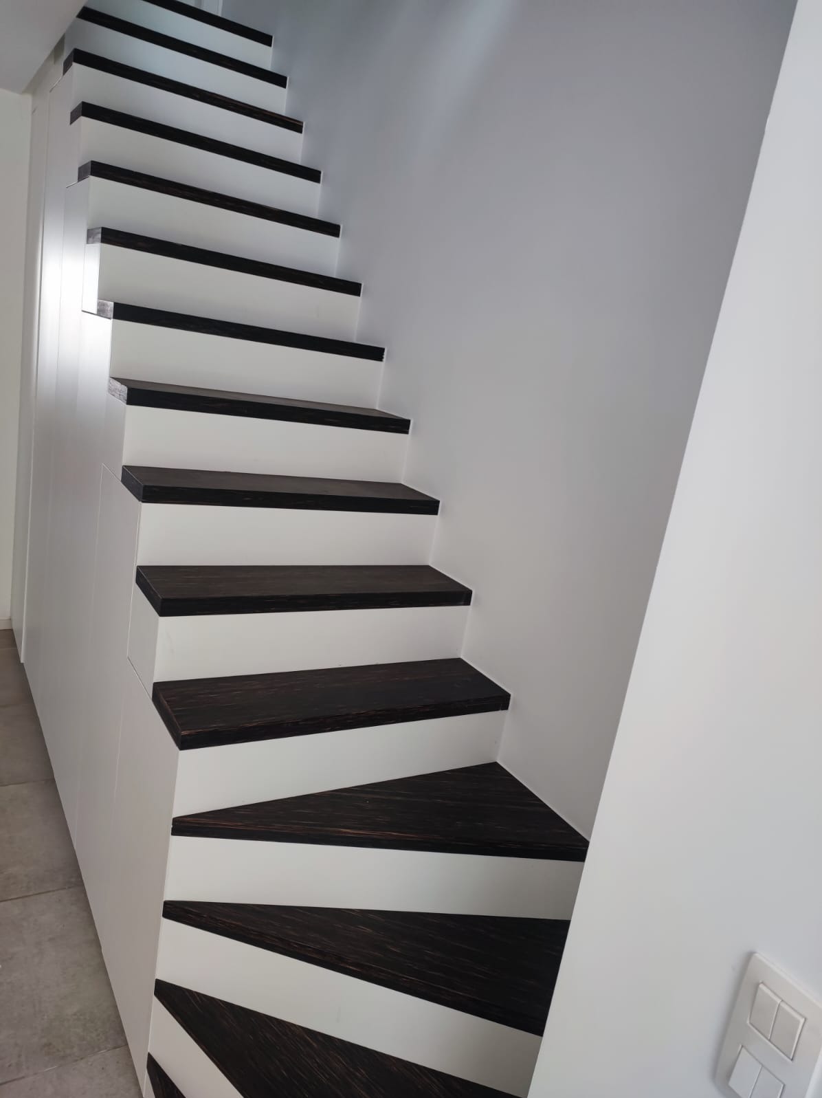 Nowoczesne schody drewniane z ciemnymi stopniami i białymi podstopnicami, widok z dołu, oświetlenie naturalne, minimalistyczny design.