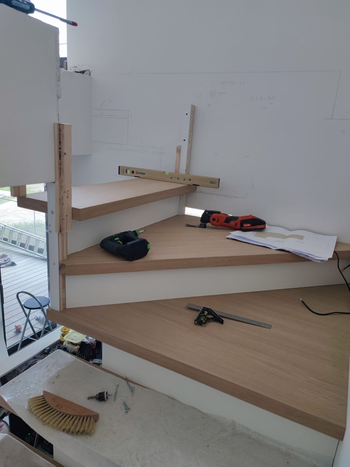 Montaż drewnianych schodów: widoczne narzędzia, poziomica, kątownik, wkrętarka. Białe ściany z zaznaczeniami, drewniane stopnie o jasnym odcieniu.