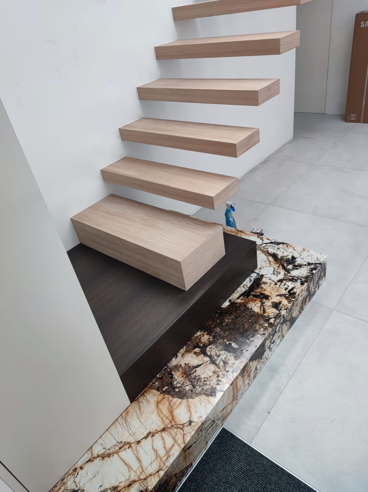 Nowoczesne schody drewniane wspornikowe z elementami wykończeniowymi z kamienia naturalnego, przy białej ścianie. Widoczne detale wykończenia i minimalistyczny design.