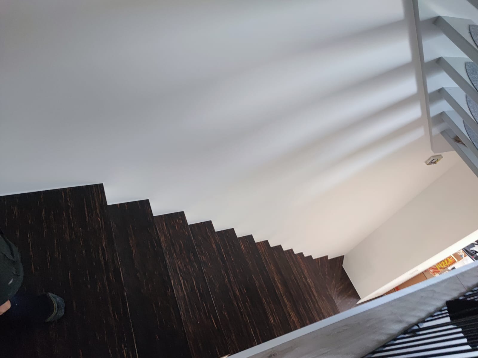 Ujęcie z góry na drewniane schody z ciemnego drewna, przy białej ścianie i suficie. Widoczny fragment osoby w dolnym lewym rogu.