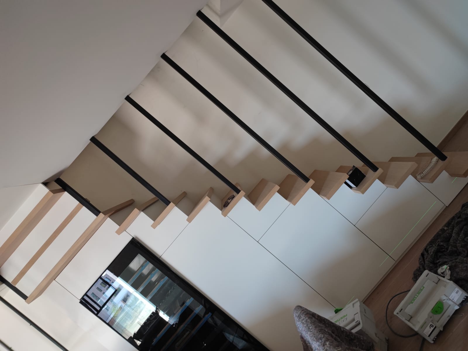 Nowoczesne schody drewniane z czarnymi balustradami i białymi panelami bocznymi, widok z góry, narzędzia w tle, jasne oświetlenie.