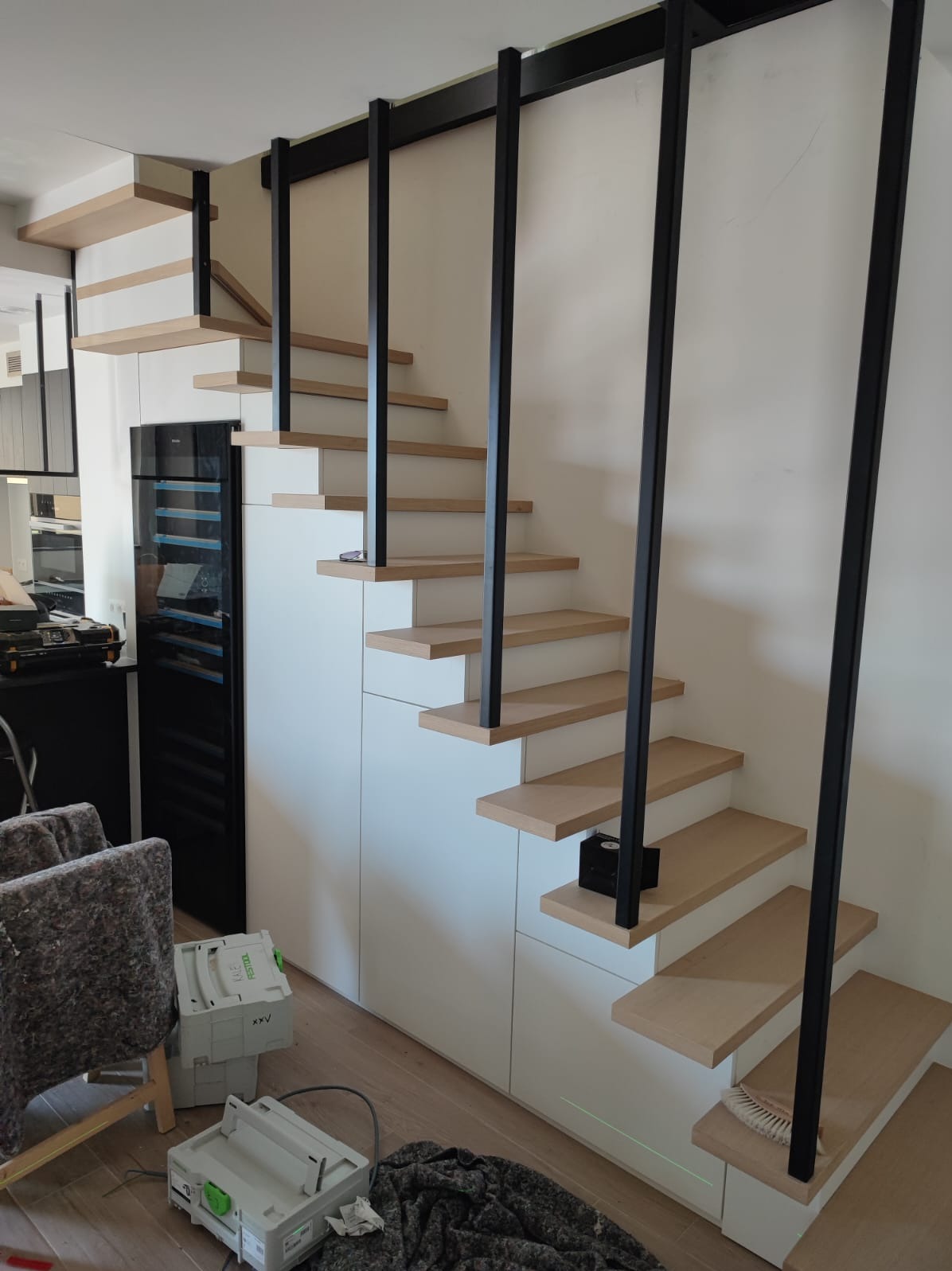 Drewniane schody z czarną, metalową balustradą w nowoczesnym wnętrzu. Widoczny fragment salonu i narzędzia pracy. Schody zintegrowane z zabudową meblową.