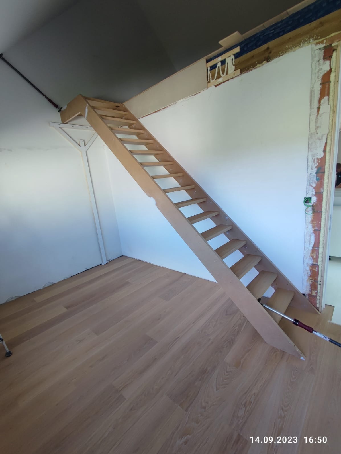 Jasne, drewniane schody strychowe zamontowane w pomieszczeniu z panelami na podłodze. Widoczne elementy konstrukcyjne i wykończeniowe.