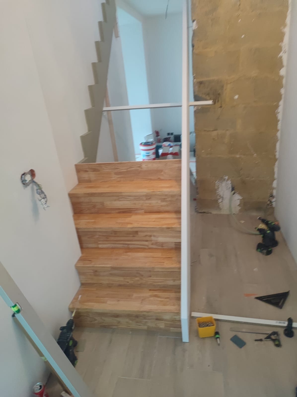 Montaż drewnianych schodów z jasnego drewna, widoczne narzędzia i fragment surowej ściany. Etap wykończeniowy, widok z dołu na pierwszy bieg schodów.