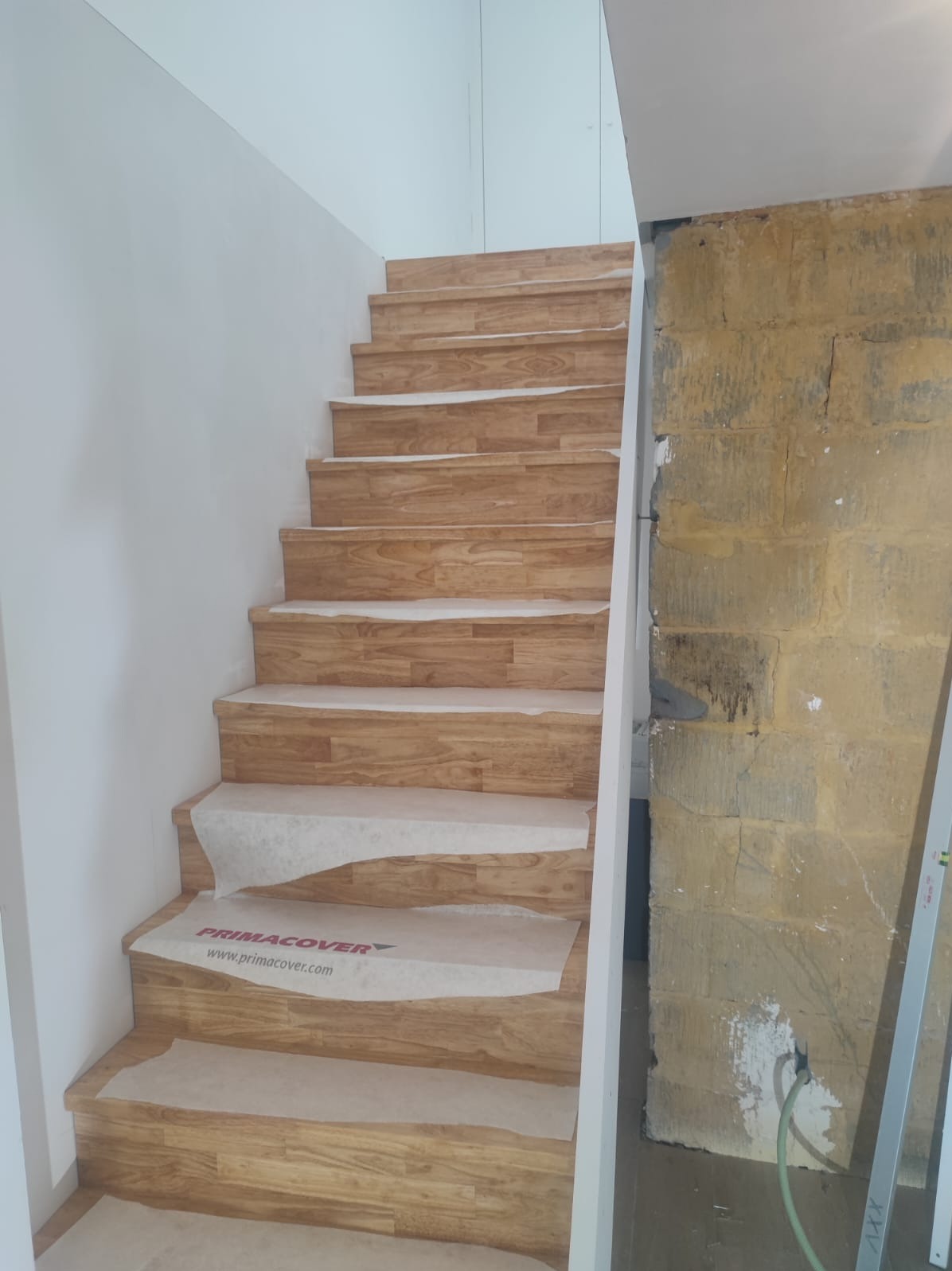 Drewniane schody z zabezpieczeniem na stopniach, jasne drewno, białe ściany, surowa ściana z kamienia. Etap prac wykończeniowych.