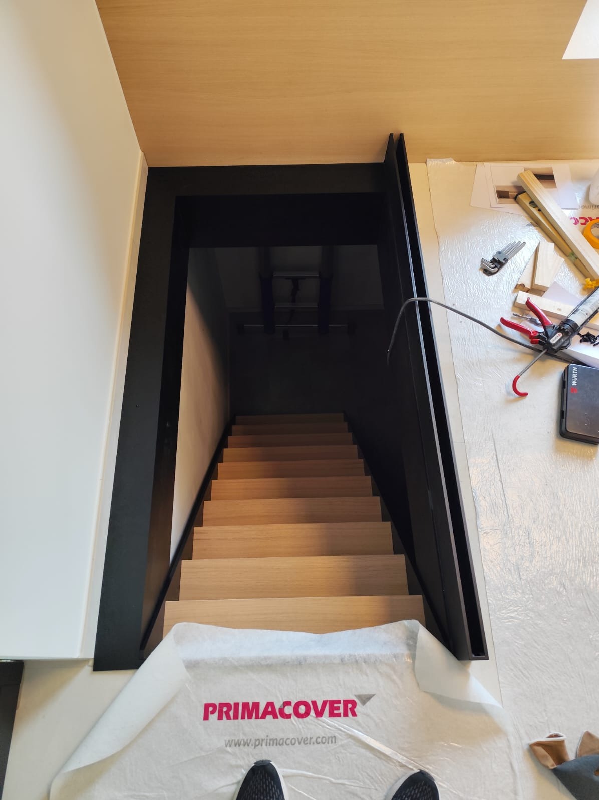 Jasne schody drewniane z czarną ramą, widok z góry, zabezpieczone folią ochronną Primacover. Częściowo widoczne narzędzia monterskie na podłodze.