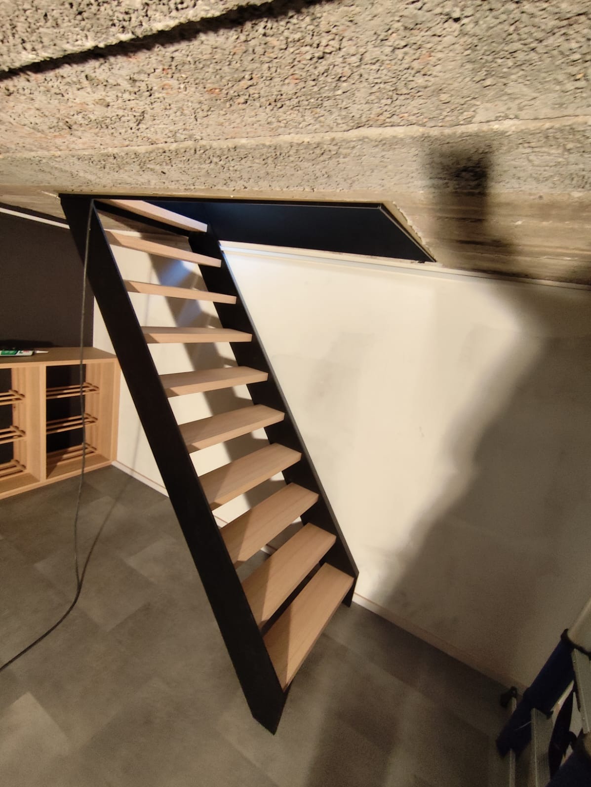 Nowoczesne schody drewniane z czarną metalową konstrukcją, prowadzące na poddasze, z widoczną surową fakturą betonowego stropu.