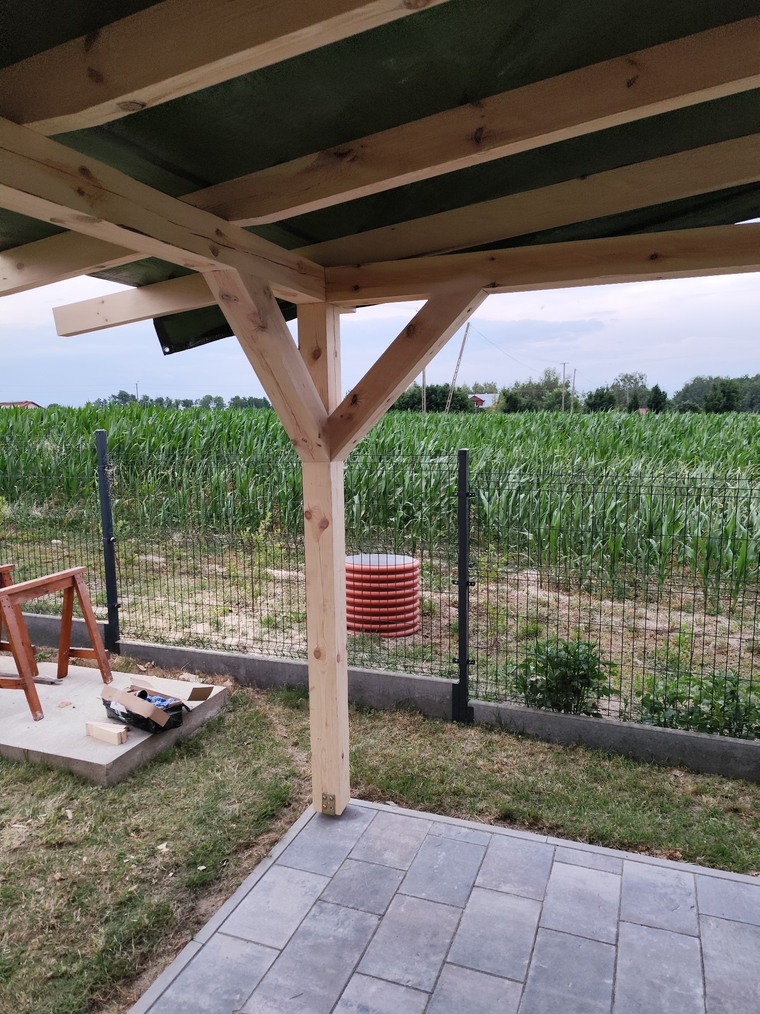 Drewniana konstrukcja altany w trakcie budowy, widoczne elementy nośne i zadaszenie. W tle pole kukurydzy za ogrodzeniem panelowym i częściowo ułożona kostka brukowa.