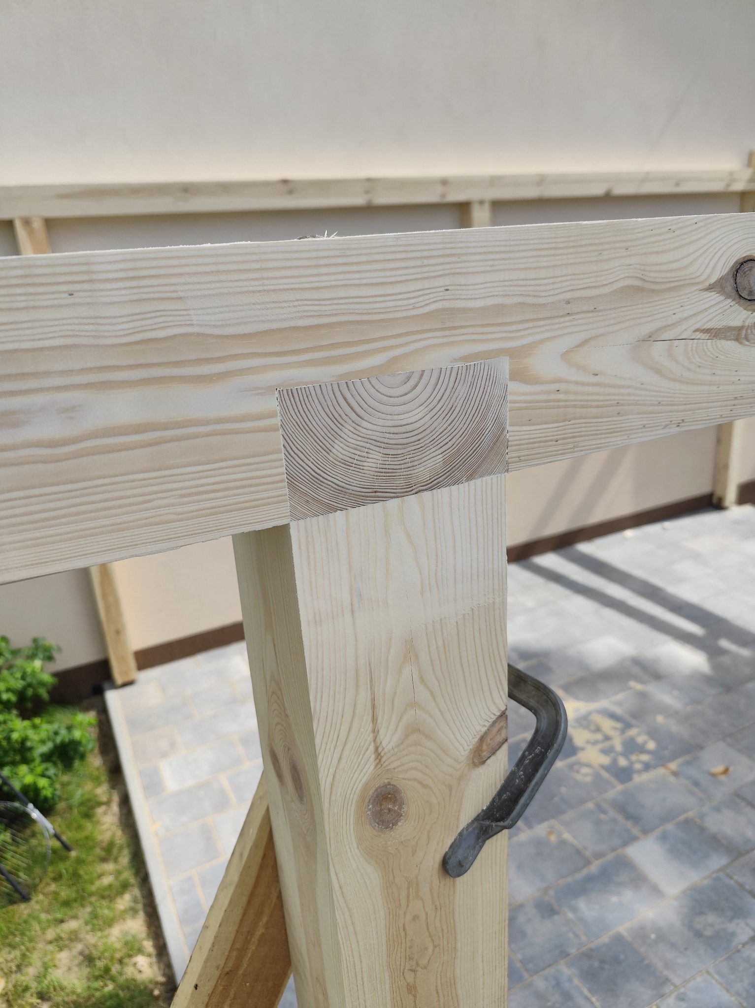 Fragment konstrukcji drewnianej, prawdopodobnie altany lub pergoli, z widocznym połączeniem belek i słupa za pomocą metalowego ścisku stolarskiego.