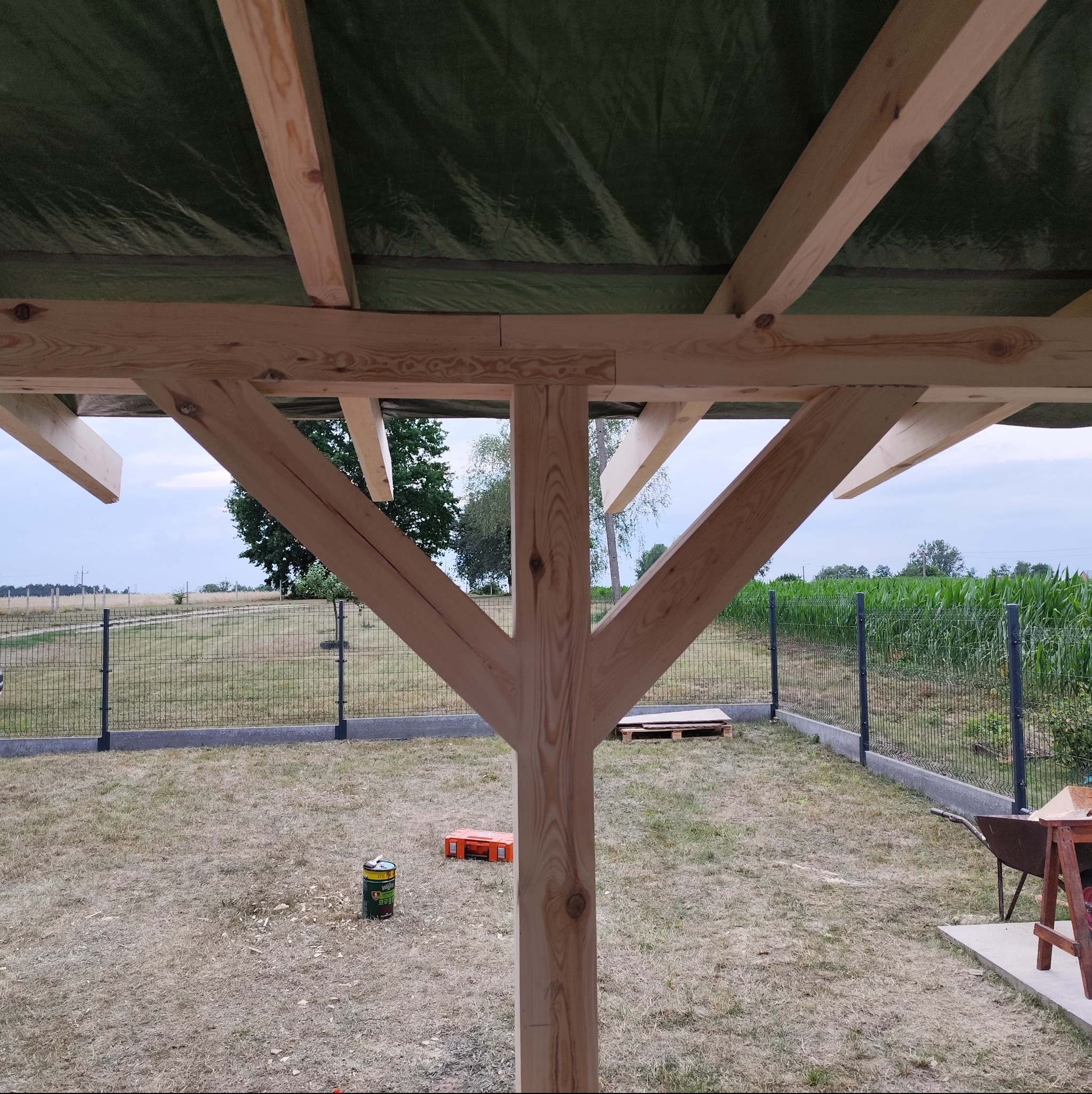 Konstrukcja drewnianej altany w trakcie budowy, widok od dołu, z widocznym zielonym zadaszeniem i elementami ogrodzenia w tle.