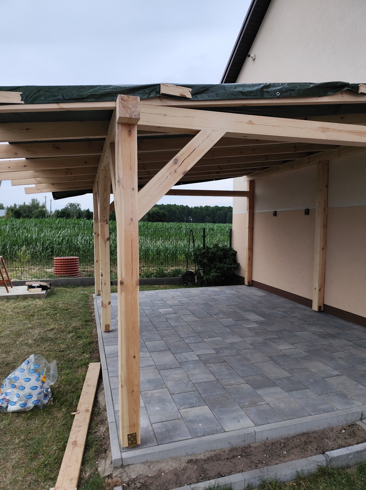 Drewniana pergola w trakcie budowy, z widocznymi elementami konstrukcyjnymi i częściowo wyłożoną kostką brukową. W tle pole kukurydzy i fragment budynku.