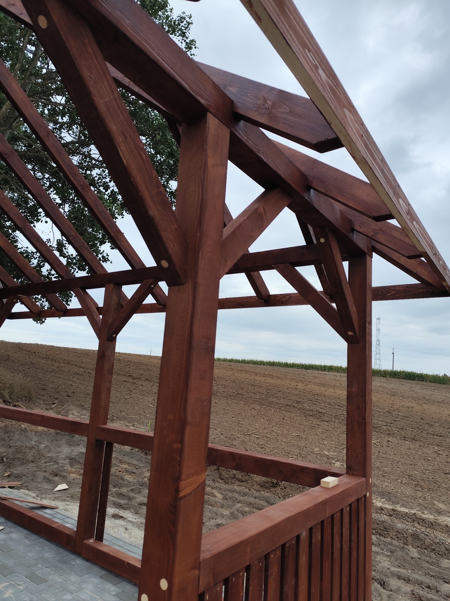 Ciemnobrązowa, drewniana konstrukcja altany w trakcie budowy, z widoczną strukturą dachu i słupami na tle zaoranego pola i pochmurnego nieba.