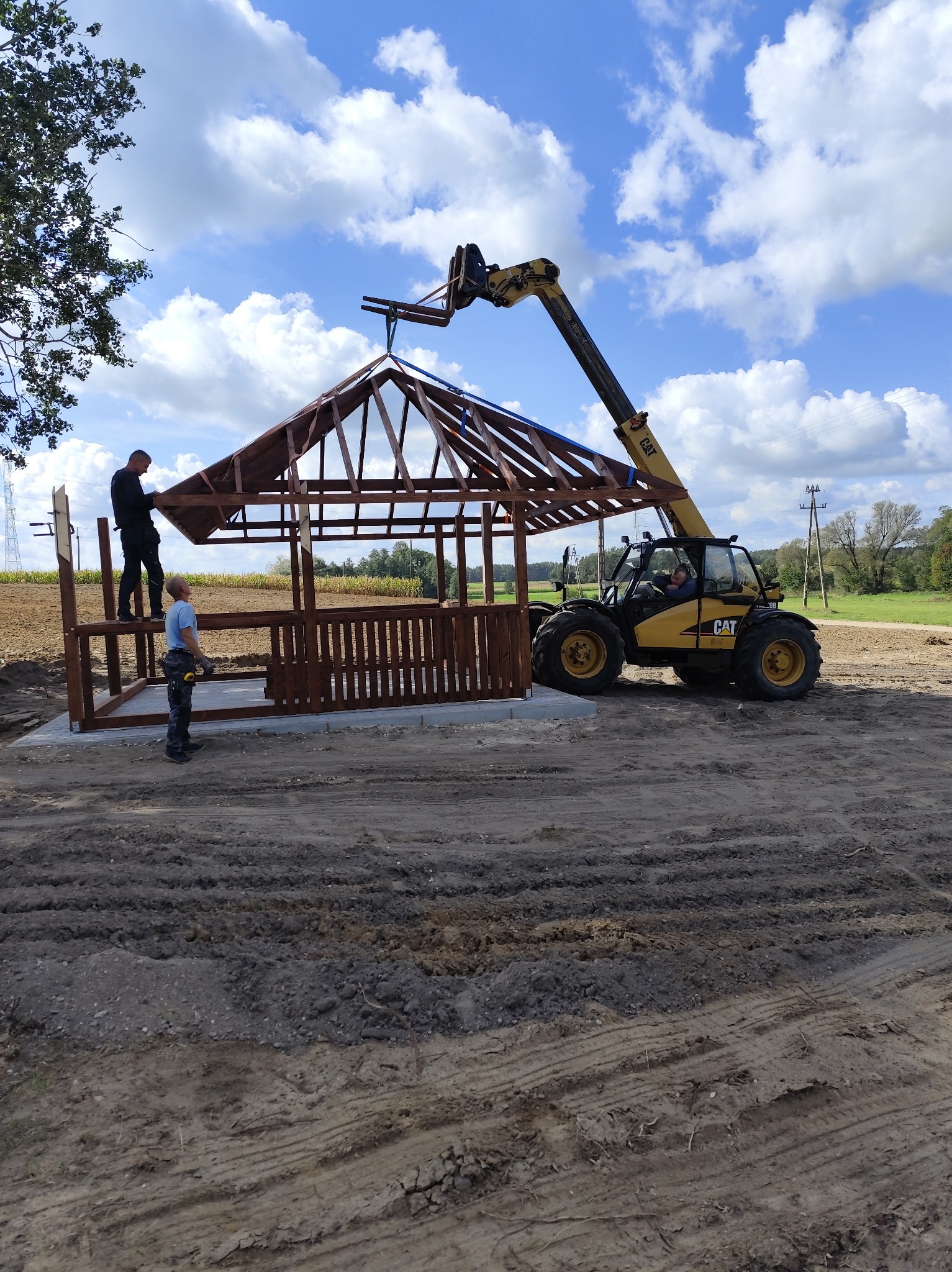 Montaż altany z drewnianą balustradą i zadaszeniem przy użyciu podnośnika CAT na tle pola i błękitnego nieba z chmurami. Dwóch monterów przy pracy.