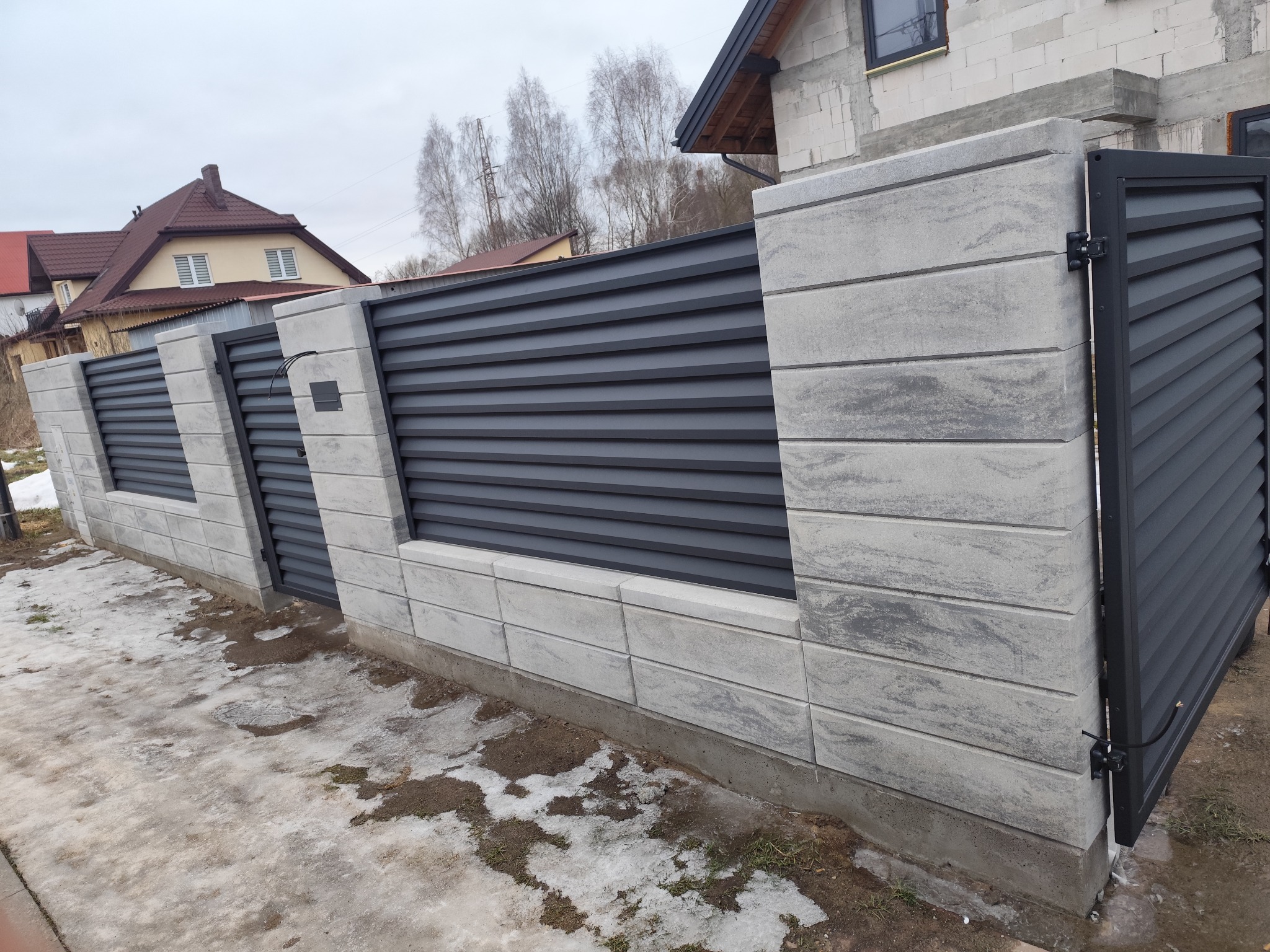 Nowoczesne ogrodzenie panelowe z betonowymi słupkami i poziomymi, grafitowymi przęsłami, częściowo pokryte śniegiem, na tle budowy domu.