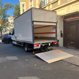 M&S-TRANSPORT SPÓŁKA Z OGRANICZONĄ ODPOWIEDZIALNOŚCIĄ - Otwarty tył białego busa z rampą rozładunkową na ulicy, zaparkowanego obok budynku z ciemnymi drzwiami wejściowymi. Widoczne zabezpieczenia wewnątrz pojazdu.