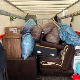 M&S-TRANSPORT SPÓŁKA Z OGRANICZONĄ ODPOWIEDZIALNOŚCIĄ - Wnętrze busa załadowane workami, meblami i sprzętem RTV. Widoczny fragment sylwetki mężczyzny w rękawicach, kontrolującego załadunek. Ściany pojazdu z szynami mocującymi.