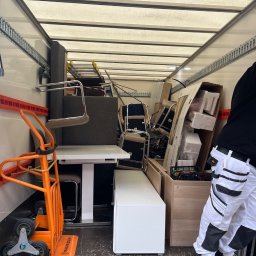 M&S-TRANSPORT SPÓŁKA Z OGRANICZONĄ ODPOWIEDZIALNOŚCIĄ - Wnętrze furgonetki wypełnionej meblami i sprzętami biurowymi podczas przeprowadzki. Widoczny wózek transportowy i fragment osoby w stroju roboczym.