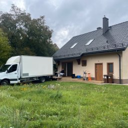 M&S-TRANSPORT SPÓŁKA Z OGRANICZONĄ ODPOWIEDZIALNOŚCIĄ - Przeprowadzka: biały samochód dostawczy zaparkowany przed domem z osobą siedzącą na platformie załadunkowej. W tle widoczny trawnik i elementy wyposażenia.