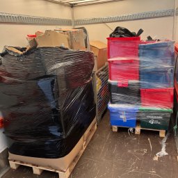 M&S-TRANSPORT SPÓŁKA Z OGRANICZONĄ ODPOWIEDZIALNOŚCIĄ - Wnętrze furgonetki z zabezpieczonym ładunkiem: duża, czarna paczka na palecie oraz stos kolorowych, foliowanych pojemników, w tym czerwone i niebieskie, gotowe do przewozu.