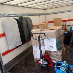 M&S-TRANSPORT SPÓŁKA Z OGRANICZONĄ ODPOWIEDZIALNOŚCIĄ - Wnętrze samochodu dostawczego z wózkiem paletowym, załadowanymi kartonami, w tym odkurzaczem Viomi, oraz wiszącymi pokrowcami na ubrania i niebieską torbą IKEA, na tle pomarańczowych szyn mocujących.