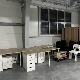 M&S-TRANSPORT SPÓŁKA Z OGRANICZONĄ ODPOWIEDZIALNOŚCIĄ - Biurka z jasnym blatem i białymi szufladami, czarne krzesła biurowe oraz regały w industrialnym wnętrzu, przygotowane do transportu lub aranżacji.