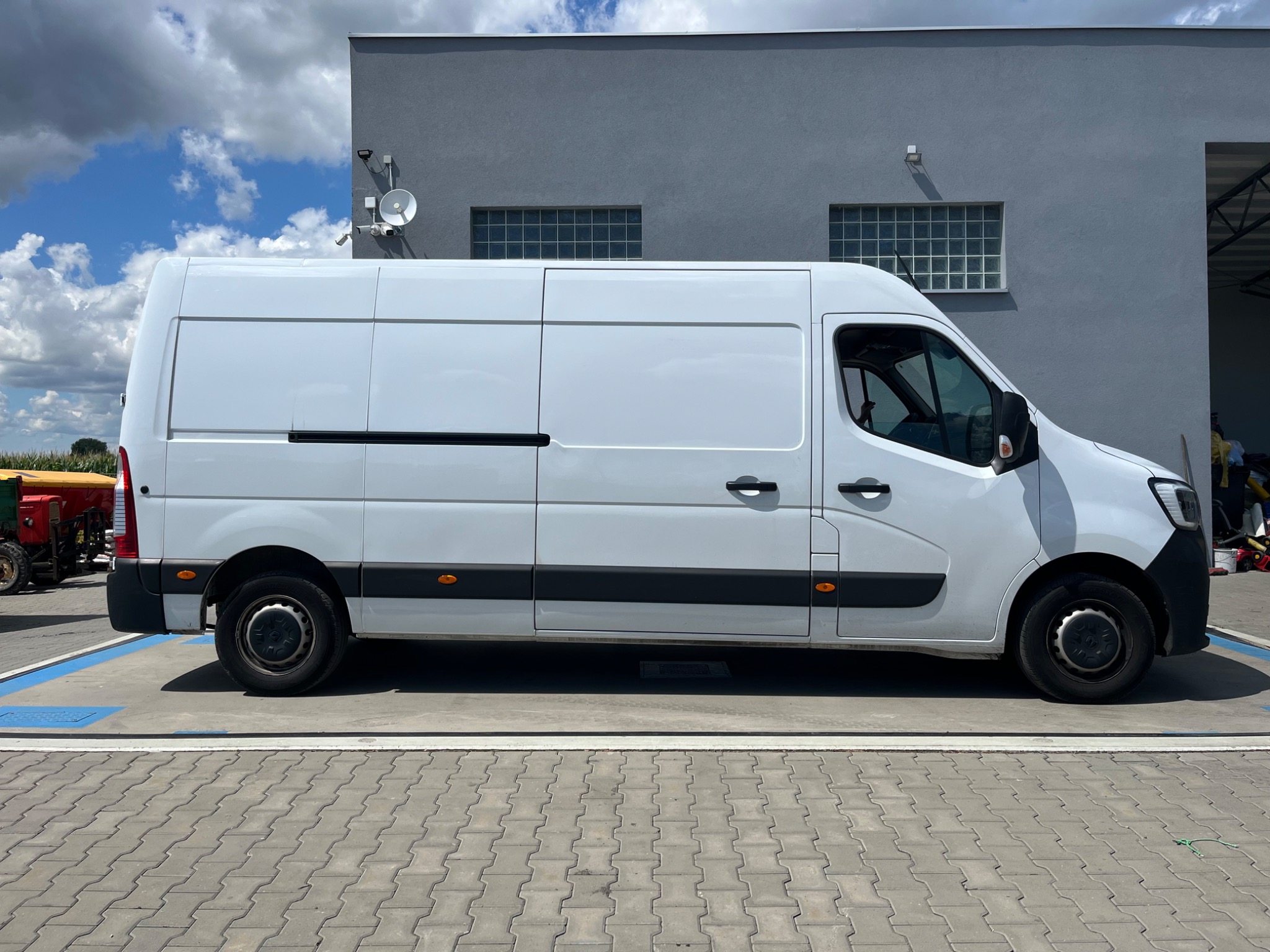 Biały furgon Renault Master na parkingu obok szarego budynku z oknami z luksferów, w tle błękitne niebo z chmurami. Widok z boku, lekki cień pod pojazdem.