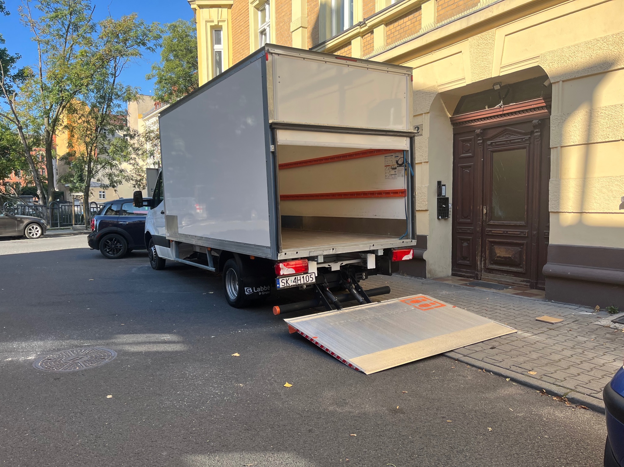 Otwarty tył białego busa z rampą rozładunkową na ulicy, zaparkowanego obok budynku z ciemnymi drzwiami wejściowymi. Widoczne zabezpieczenia wewnątrz pojazdu.