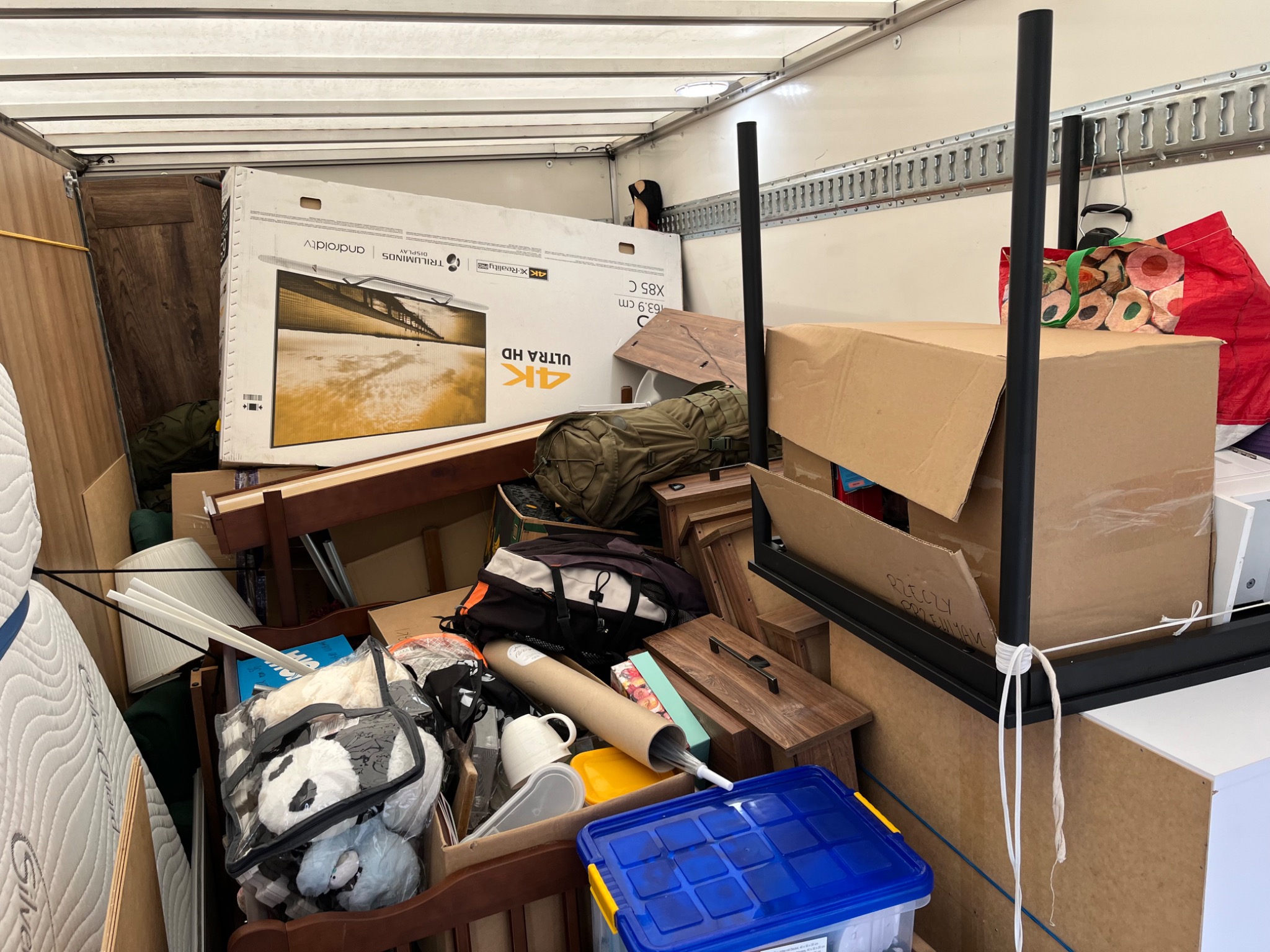 Wnętrze furgonetki załadowane różnorodnymi przedmiotami podczas przeprowadzki, w tym pudełko z telewizorem 4K, meble, torby i inne rzeczy osobiste, zabezpieczone sznurkami.