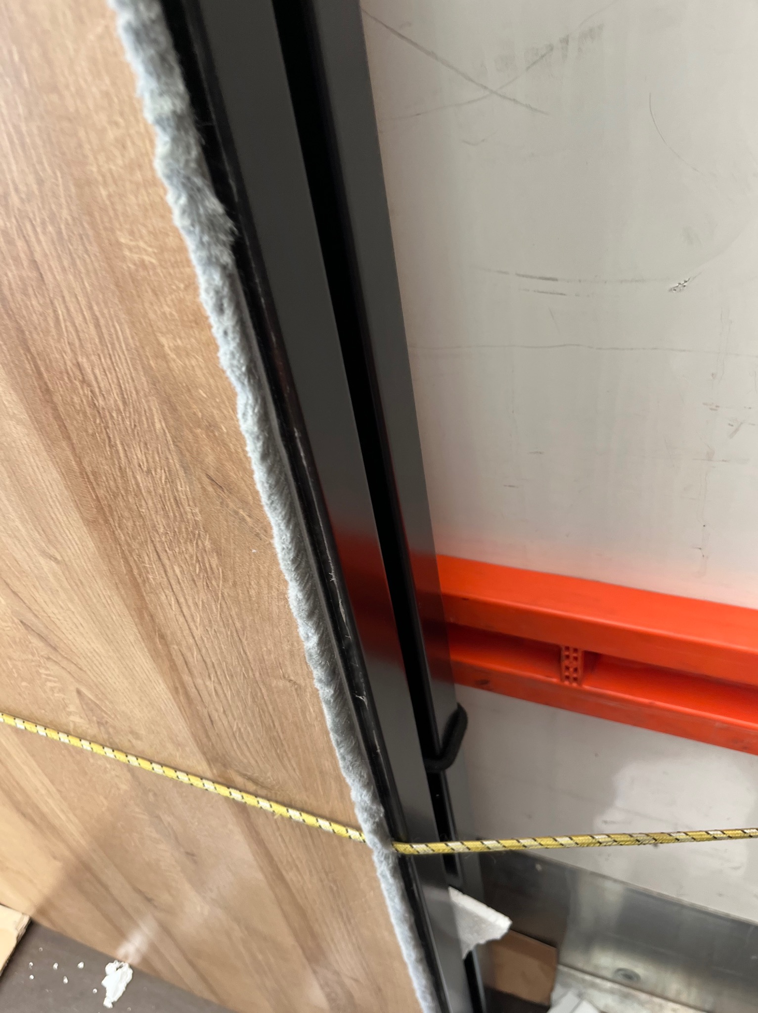 Fragment zabezpieczonej płyty meblowej z szarą uszczelką i czarną ramą, zabezpieczonej żółtą liną transportową na tle regału magazynowego.