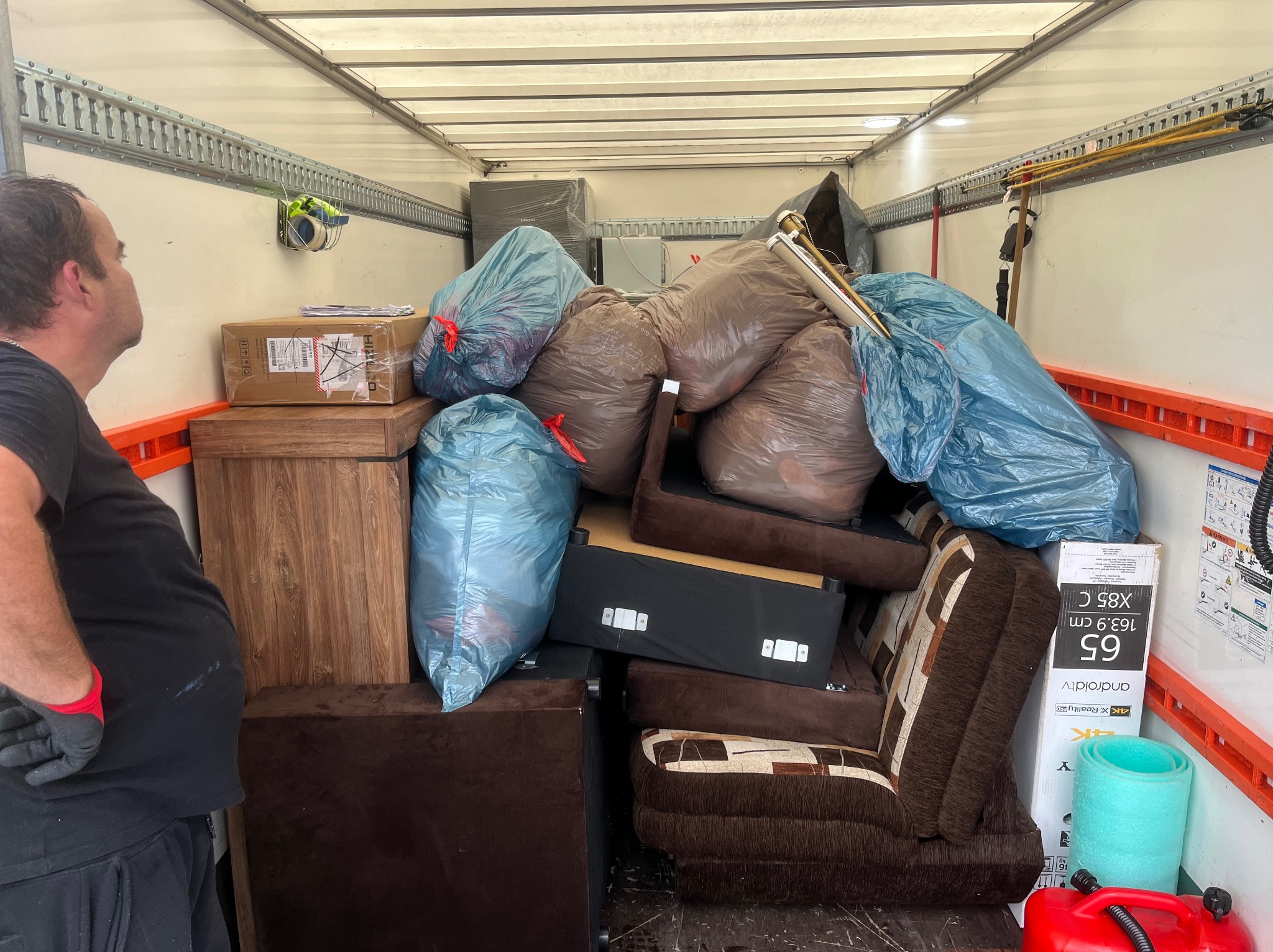 Wnętrze busa załadowane workami, meblami i sprzętem RTV. Widoczny fragment sylwetki mężczyzny w rękawicach, kontrolującego załadunek. Ściany pojazdu z szynami mocującymi.